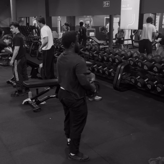Une salle de sport bondée : Pious, en haut rouge, parmi des gens qui s'entraînent près des racks d'haltères.