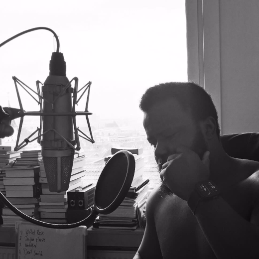 En noir et blanc : Pious est assis près d'un micro de studio, le menton dans la main, à côté d'une fenêtre lumineuse.